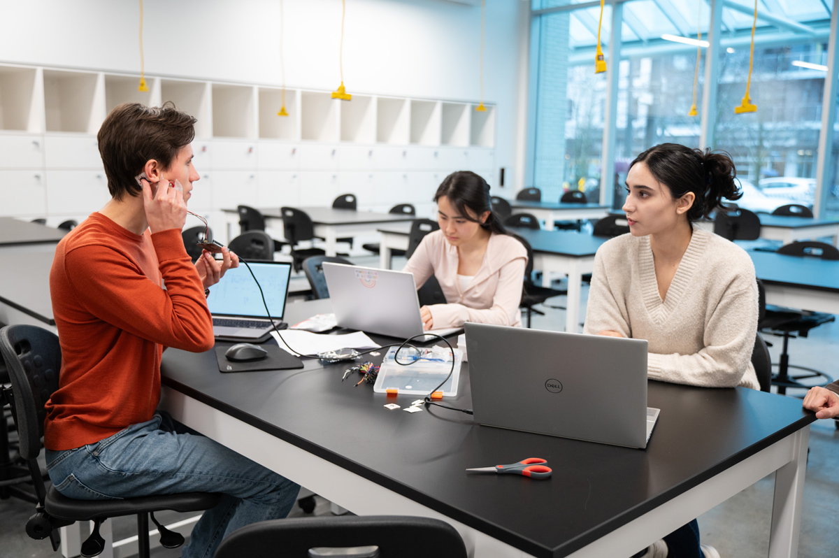 Three students working on laptops at a table. One is holding a device to his head.