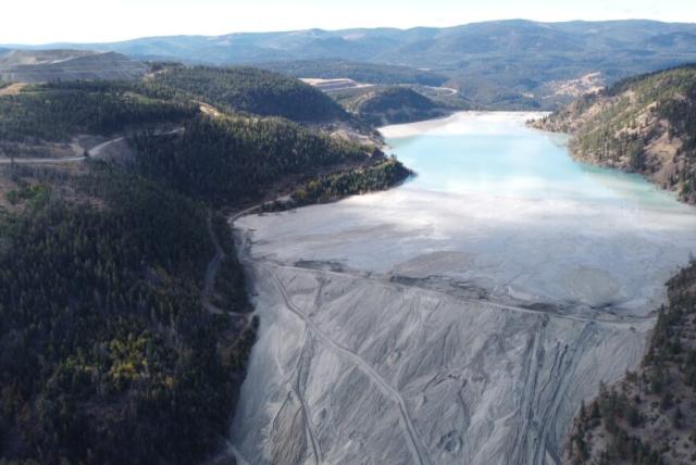 The steep wall of a massive tailings dam holds back cloudy water between treed hills