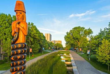 A tall totem pole featuring a human figure holding a serpant that wraps around them, stands at the entry to a long tree-lined boulevard.