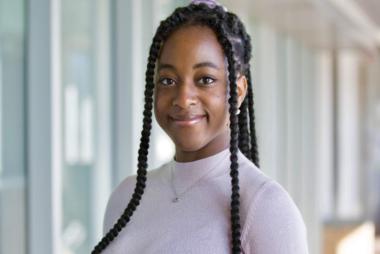 UBCO Electrical Engineering student Aliyah Ayorinde smiling at the camera. She is centred outdoors as a panel of building windows frames her on the left.