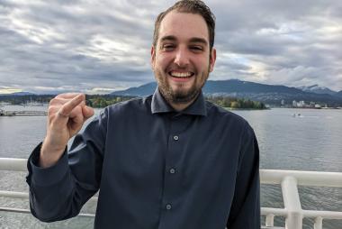UBC Integrated Engineering student Mark Greenwood smiles at the camera while holding his pinky up to showcase his iron ring. He is centred and posed in front of a body of water.