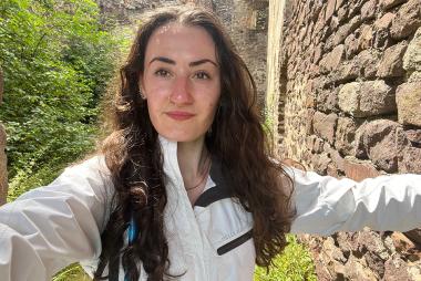 Woman with long dark hair in a white coat takes a selfie with greenery and stone in the background
